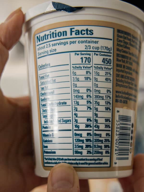 Close-up of a hand holding a container, focusing on the nutrition facts label. It lists calories, fats, cholesterol, sodium, carbohydrates, protein, and vitamins, with values per serving and per container. A thumb is visible at the bottom.